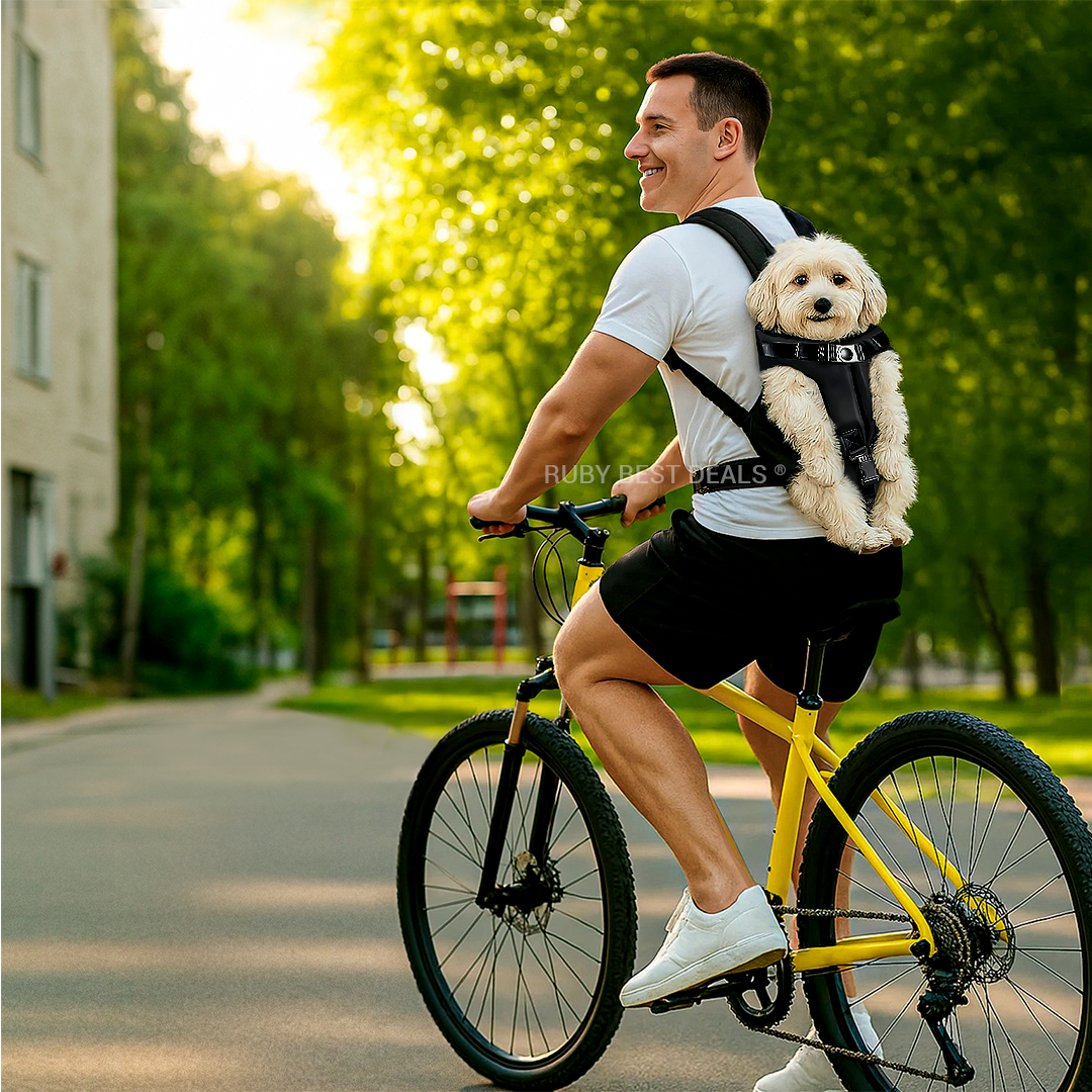 Man riding a bike with a small dog in a black backpack carrier on his back, surrounded by green trees
