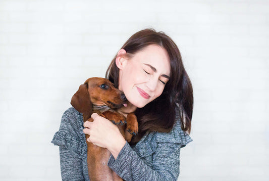 A woman with closed eyes and a smile, hugging a brown dachshund puppy close to her face.