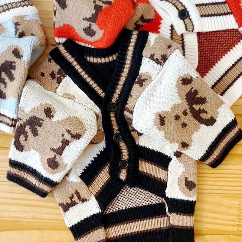 Close-up of a beige dog cardigan with a teddy bear pattern, featuring black buttons and beige trim, laid on a wooden surface