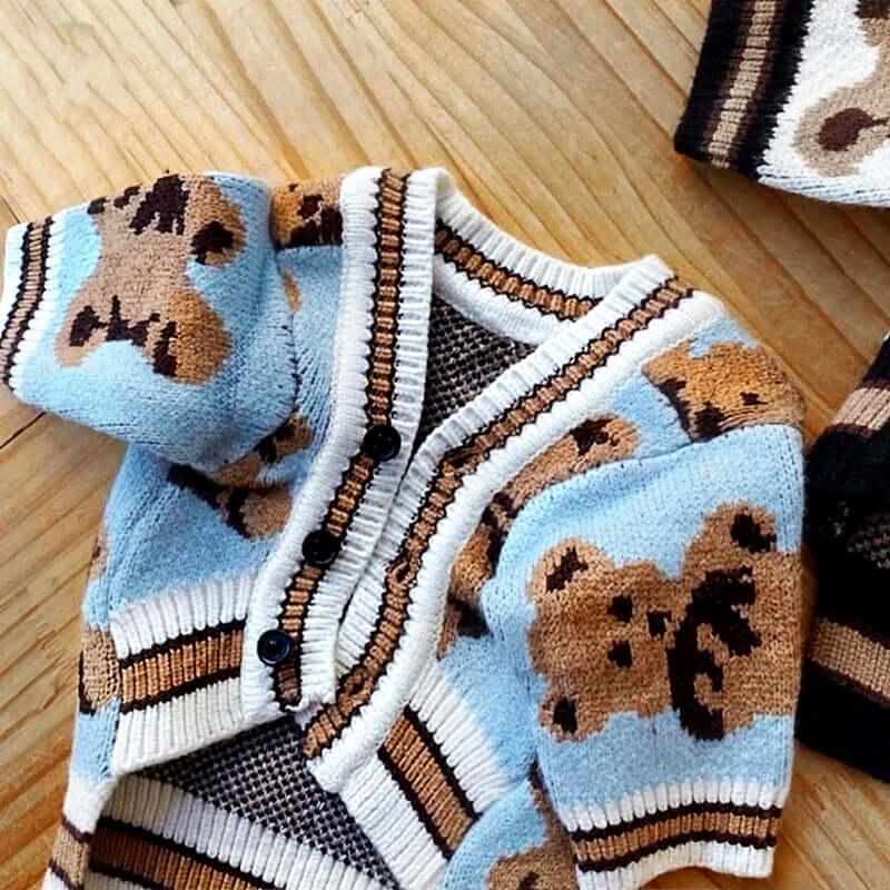 Close-up of a light blue dog cardigan with a teddy bear pattern, featuring black buttons and beige trim, laid on a wooden surface