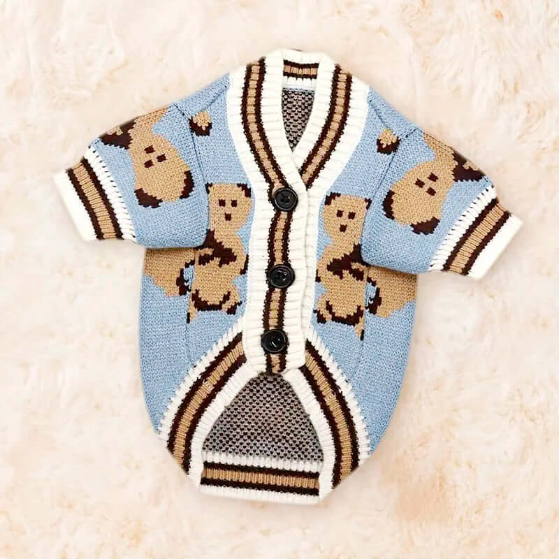 A blue dog cardigan with black trim, black buttons, and a teddy bear pattern, laid flat on a soft, cream-colored surface