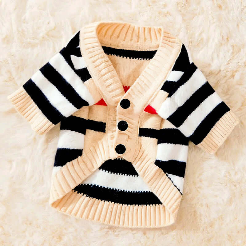Beige dog cardigan with black and white stripes, featuring three black buttons down the front, lying on a cream-colored surface