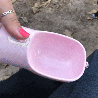 A person is holding a pink PetPal Water & Snack Bottle with a scoop shape, pouring water into it for a dog to drink from.