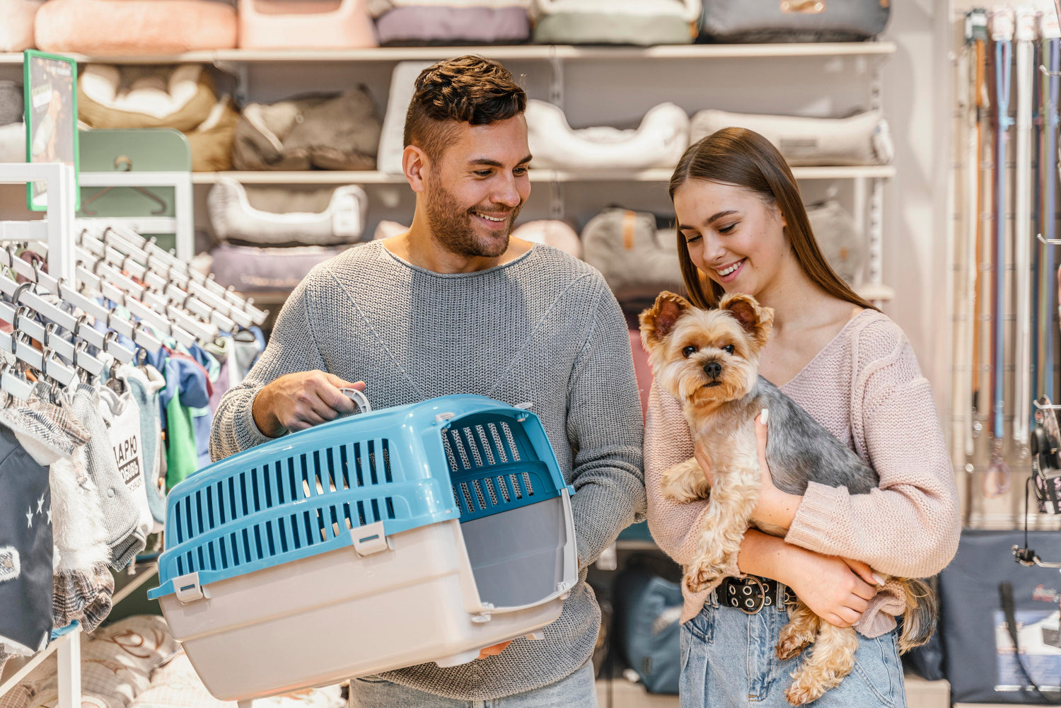 Couple in pet store: man holds blue carrier, woman holds small dog. Shelves stocked with pet supplies.